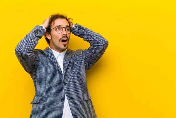 young handsome man with open mouth, looking horrified and shocked because of a terrible mistake, raising hands to head against orange wall