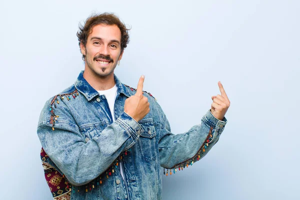 young handsome man smiling happily and pointing to side and upwards with both hands showing object in copy space isolated against flat wall