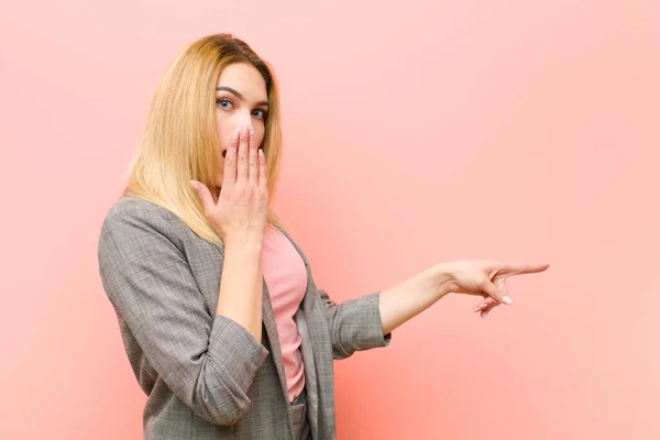 young pretty blonde woman feeling happy, shocked and surprised, covering mouth with hand and pointing to lateral copy space against flat wall