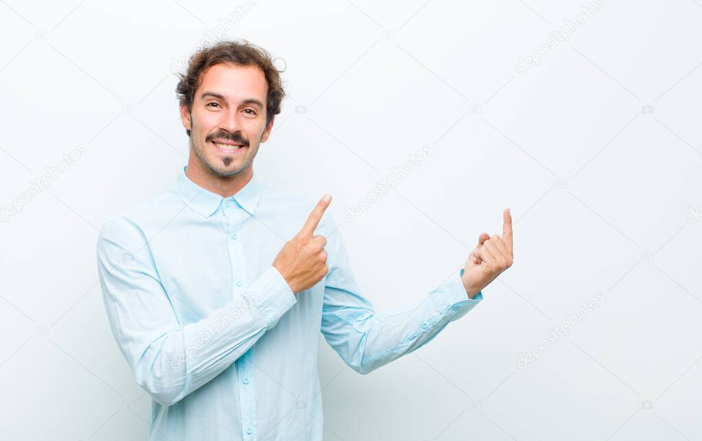 young handsome man smiling happily and pointing to side and upwards with both hands showing object in copy space against white wall