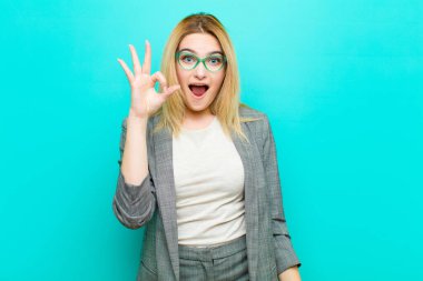 Genç, güzel sarışın kadın kendini başarılı ve tatmin olmuş hissediyor, ağzı açık gülümsüyor, düz duvara el işareti yapıyor.