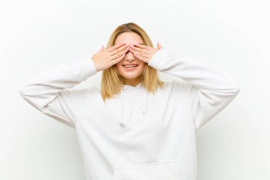 Genç sarışın kadın gülümsüyor ve mutlu hissediyor, iki eliyle gözlerini kapatıyor ve beyaz duvara karşı inanılmaz bir sürpriz bekliyor.