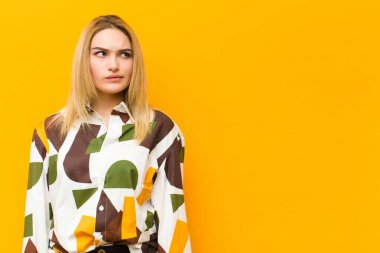 Şaşkın ve kafası karışmış genç sarışın kadın, bir sorunu çözmeye çalışıyor ya da turuncu duvara karşı düşünüyor.