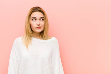 Genç, güzel sarışın kadın kafası karışmış ve şaşkın görünüyor. Bir sorunu çözmeye çalışıyor ya da pembe düz duvara karşı düşünüyor.