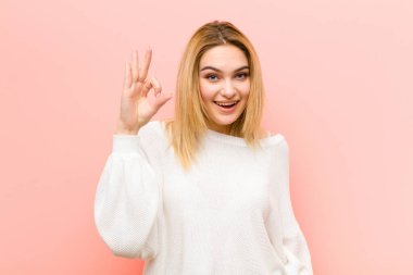 Genç güzel sarışın kadın kendini başarılı ve tatmin olmuş hissediyor, ağzı açık gülümsüyor, pembe düz duvara karşı elle işaret veriyor.