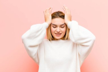 Genç ve güzel sarışın kadın stresli ve endişeli, depresif ve hayal kırıklığına uğramış hissediyor, iki elini de pembe düz duvara doğru kaldırıyor.