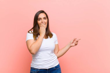 Genç, güzel İspanyol kadın mutlu, şok olmuş ve şaşırmış hissediyor. Ağzını eliyle kapatıyor ve pembe duvara karşı yanal fotokopi alanını işaret ediyor.