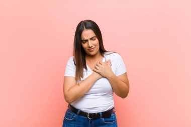 Genç, güzel İspanyol bir kadın mutlu, arkadaş canlısı ve pozitif hissediyor, gülümsüyor ve pembe duvara yaslanmış ellerle bir portre veya fotoğraf çerçevesi yapıyor.