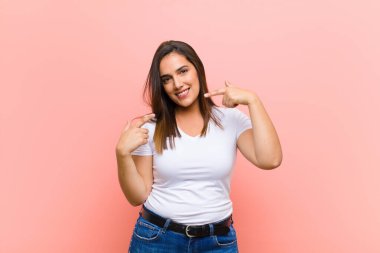 Genç, güzel İspanyol kadın, kendine güvenerek gülümsüyor. Pembe duvara karşı pozitif, rahat, tatmin olmuş bir tavır sergiliyor.