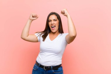 Genç, güzel İspanyol bir kadın muzaffer bir şekilde bağırıyor. Heyecanlı, mutlu ve şaşırmış görünüyor. Pembe duvara karşı kutlama yapıyor.