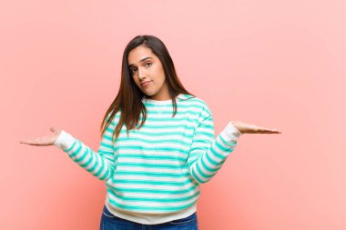 Genç, güzel, İspanyol bir kadın kafası karışık ve kafası karışık. Doğru cevap veya karardan emin değil. Pembe duvara karşı bir seçim yapmaya çalışıyor.