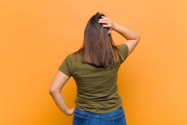 Genç güzel bir kadın düşünüyor ya da şüphe ediyor, kafasını kaşıyor, şaşkın ve şaşkın hissediyor, turuncu duvara yaslanmış arka ya da arka pencereden bakıyor.