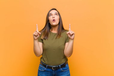 Şok olmuş, şaşırmış ve ağzı açık genç bir kadın, iki eliyle yukarıya doğru bakıyor turuncu duvara izole edilmiş alanı kopyalamak için.