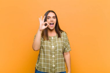 Genç, güzel bir kadın kendini başarılı ve tatmin olmuş hissediyor. Ağzı açık gülümsüyor. Elleriyle turuncu duvara yaslanmış bir işaret veriyor.