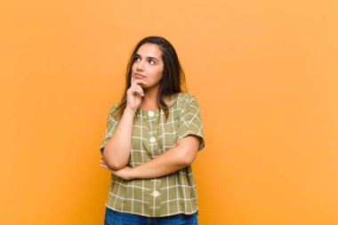 young pretty woman with a concentrated look, wondering with a doubtful expression, looking up and to the side isolated against orange wall clipart
