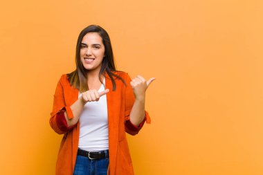 Genç, güzel bir kadın neşeli ve tesadüfen fotokopi alanını işaret ederek turuncu duvara yaslanmış mutlu ve tatmin olmuş hissediyor.