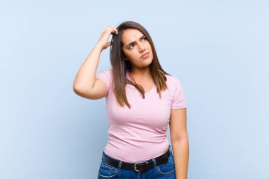 Genç, güzel bir kadın şaşkın ve kafası karışmış hissediyor. Kafasını kaşıyor ve mavi arka plana bakıyor.