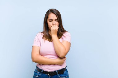 Genç, güzel bir kadın ciddi, düşünceli ve endişeli, elleri çenesine değmiş, mavi arka planda yan bakıyor.