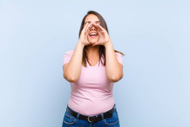 Genç, güzel bir kadın mutlu, heyecanlı ve pozitif hissediyor. Elleri ağızda, mavi arka planda bağırıyor.