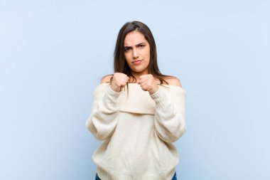 Kendine güvenen, sinirli, güçlü ve agresif görünen genç bir kadın. Yumrukları boks pozisyonunda mavi arka planda dövüşmeye hazır.