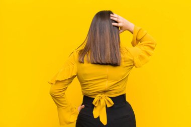 Genç güzel bir kadın düşünüyor ya da şüphe ediyor, kafasını kaşıyor, şaşkın ve şaşkın hissediyor, turuncu duvara yaslanmış arka ya da arka pencereden bakıyor.