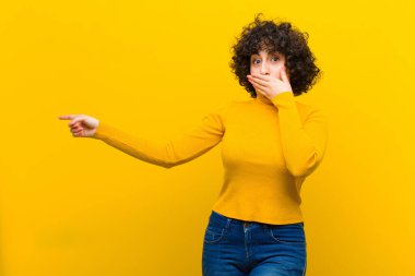 Genç, güzel afro kadın kendini mutlu, şok olmuş ve şaşırmış hissediyor. Ağzını eliyle kapatıyor ve yanal fotokopi alanını işaret ediyor.