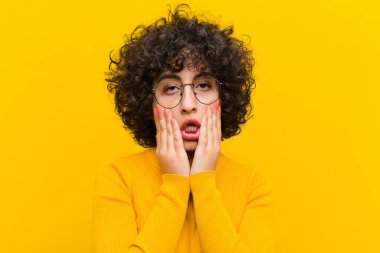 Genç, güzel afro kadın şok olmuş ve korkmuş hissediyor, ağzı açık ve elleri yanaklarında korkmuş görünüyor.