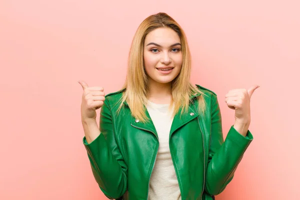 若いです可愛いですブロンド女性笑顔楽しく見て幸せ 気楽な気持ちと正の両方の親指アップでピンクフラット壁に対して — ストック写真