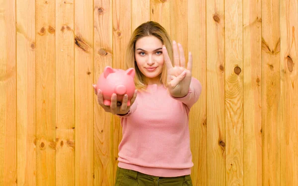 年轻漂亮的金发女人 靠着木墙有一个小猪银行 — 图库照片