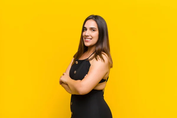 young pretty woman smiling to camera with crossed arms and a happy, confident, satisfied expression, lateral view isolated against orange wall