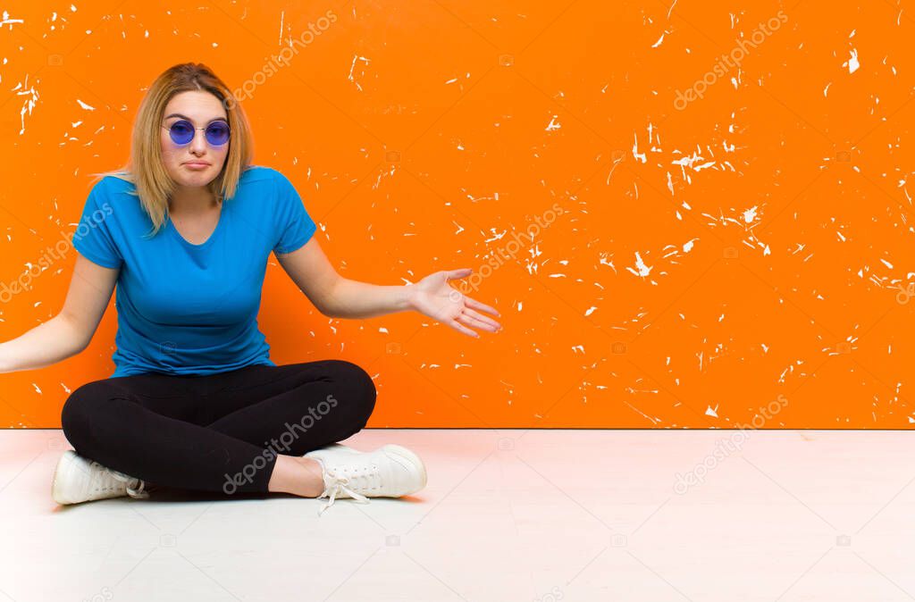 young blonde woman feeling clueless and confused, having no idea, absolutely puzzled with a dumb or foolish look sitting on the floor
