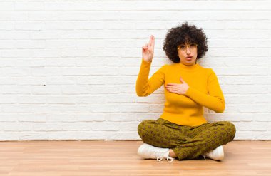 Genç, güzel afro kadın mutlu, kendinden emin ve güvenilir görünüyor, gülümsüyor ve zafer işareti gösteriyor, olumlu bir tavırla yerde oturuyor.