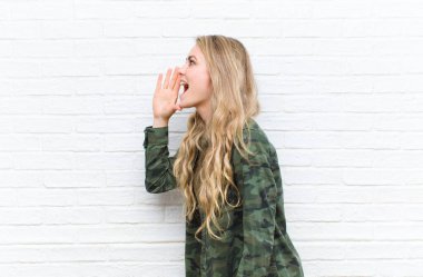 Genç sarışın kadın profil görüntüsü, mutlu ve heyecanlı görünüyor, bağırıyor ve duvarın arka planındaki boşluğu kopyalamak için çağırıyor.