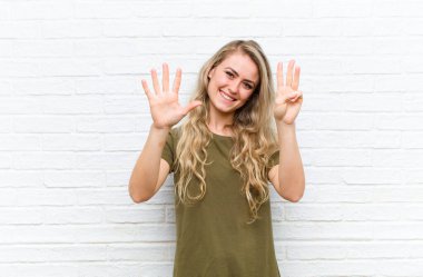 Genç sarışın kadın gülümsüyor ve arkadaş canlısı görünüyor, 8 ya da 8 numarayı gösteriyor, elleri önde, tuğla duvar arkasında geriye doğru sayıyor.