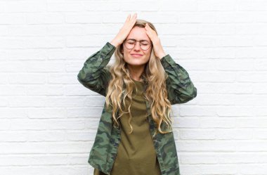 Genç sarışın kadın stresli ve endişeli, depresyonda ve baş ağrısı yüzünden hayal kırıklığına uğramış hissediyor, iki elini de duvara yaslıyor.