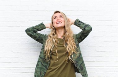 Genç sarışın kadın gülümsüyor ve rahatlıyor, tatmin oluyor, kaygısız gülüyor ve tuğla duvarlara yaslanıyor.