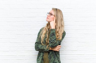 Yoğunlaşmış bakışlı genç sarışın kadın, şüpheli bir ifadeyle, yukarı ve yana bakıyor tuğla duvar arka planı karşı