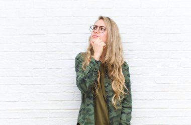 Genç sarışın kadın düşünüyor, şüpheci ve şaşkın hissediyor, farklı seçeneklerle, tuğla duvar arka planına karşı hangi kararı vereceğini merak ediyor.