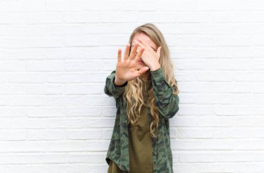 Genç sarışın kadın yüzünü eliyle kapatıyor ve kamerayı durdurmak için diğer elini öne koyuyor. Duvara dayalı fotoğraf ve resimleri reddediyor.