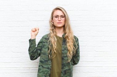 Genç sarışın kadın kendini ciddi, güçlü ve asi hissediyor, yumruğunu kaldırıyor, tuğla duvar arkasında devrim için protesto ediyor ya da savaşıyor.