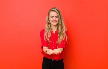 Genç sarışın kadın mutlu bir şekilde gülümsüyor, kendine güvenen, pozitif bakışlı, kırmızı duvara karşı bir nesne ya da konsept sunuyor ve gösteriyor.