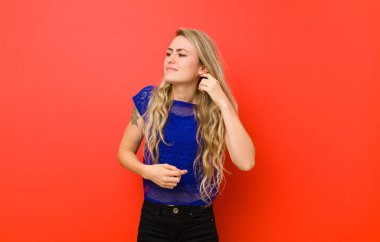 Genç sarışın kadın stresli, hayal kırıklığına uğramış ve yorgun hissediyor, boynunu ovuyor, endişeli ve sorunlu bir şekilde kırmızı duvara bakıyor.