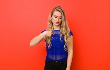 Genç sarışın kadın neşeyle ve gelişigüzel gülümsüyor, aşağıya bakıyor ve göğsünü kırmızı duvara gösteriyor.