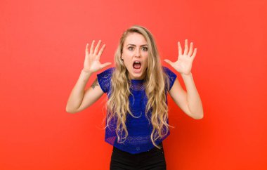 Genç sarışın kadın panik ya da öfke içinde bağırıyor, şok olmuş, korkmuş ya da öfkeli, elleri kırmızı duvara dayalı.