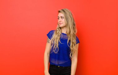 Genç sarışın kadın merak ediyor, mutlu düşünceler ve fikirler düşünüyor, hayal kuruyor, kırmızı duvara karşı uzayı kopyalamaya çalışıyor.