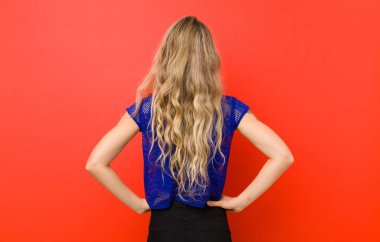 Genç sarışın kadın kafası karışık ya da dolgun hissediyor ya da şüphe ve sorular, merak ediyor, elleri kalçasında, kırmızı duvarda dikiz aynasıyla