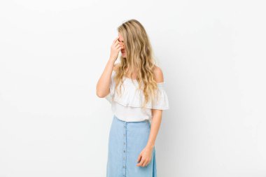 Genç sarışın kadın stresli, mutsuz ve hayal kırıklığına uğramış, alnına dokunuyor ve beyaz duvara karşı şiddetli baş ağrısı çekiyor.