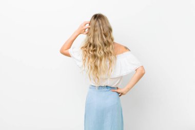 Sarışın genç kadın kendini cahil ve kafası karışık hissediyor, bir çözüm düşünüyor, eli kalçasında, diğeri kafasında, beyaz duvara karşı dikiz aynasından bakıyor.