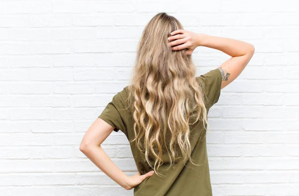 young blonde woman thinking or doubting, scratching head, feeling puzzled and confused, back or rear view against brick wall background