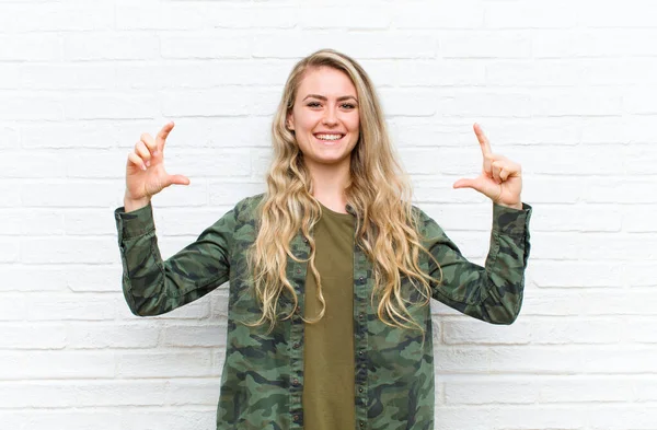 young blonde woman framing or outlining own smile with both hands, looking positive and happy, wellness concept against brick wall background